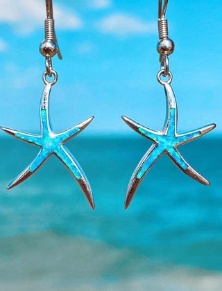 Blue opal starfish drop earrings in silver tone, photographed against the sea