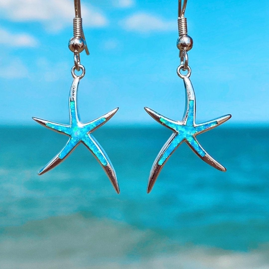 Blue opal starfish drop earrings in silver tone, photographed against the sea