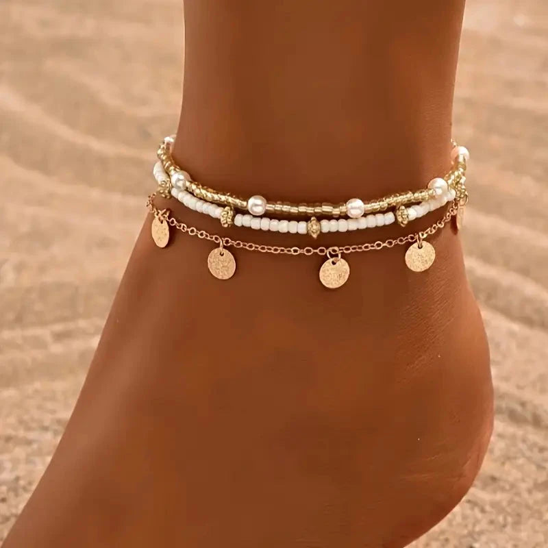 Layered gold-tone anklets worn together on an ankle on the sand
