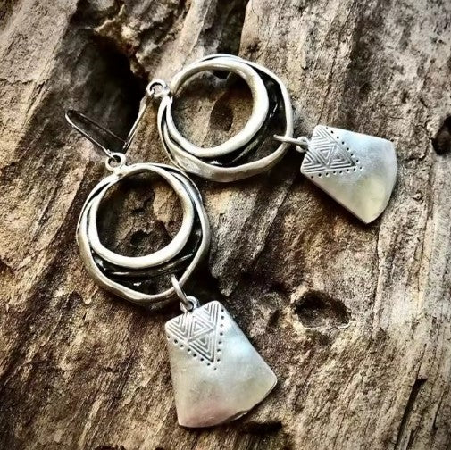Silver-tone dangle earrings with a hammered open ring and a stamped shield-shaped charm, photographed on weathered wood
